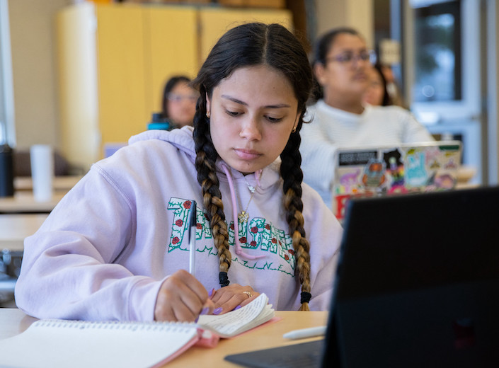 Child Development Early Childhood Development student writes in a notebook in her classroom. She is of Latinx descent and has thick dark braids and a nose ring.