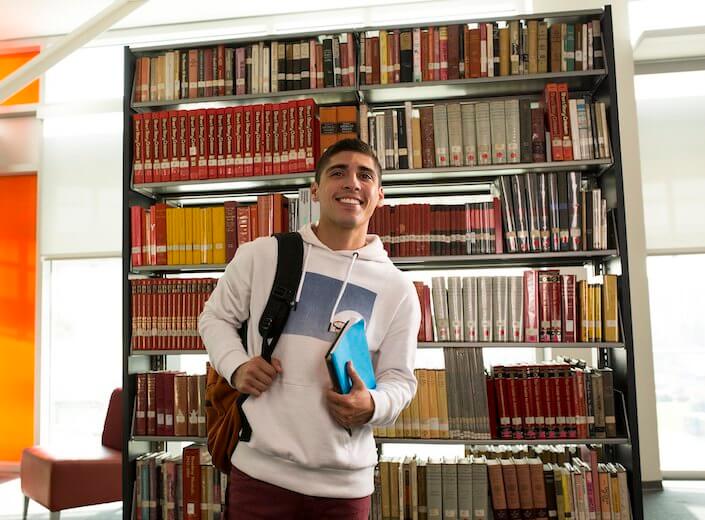 Boy in library Latino college student in white hoodie holds a book and has backpack slung over his shoulder. He is standing in front of library stacks.