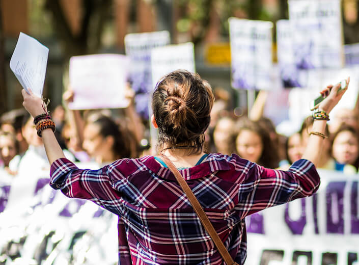 Women's March Young woman with light brown hair pulled up into a messy bun wears a red flannel and has both arms outstreched facing an out-of-focus crowd of protesters holdig signs.