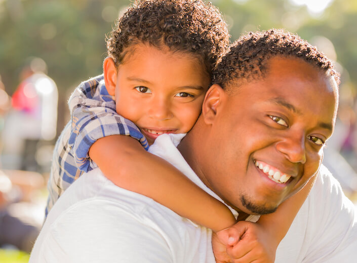 Dad and son. African-American dad and son. The young son throws his arms around dad's neck. They are outside in a sunny park.