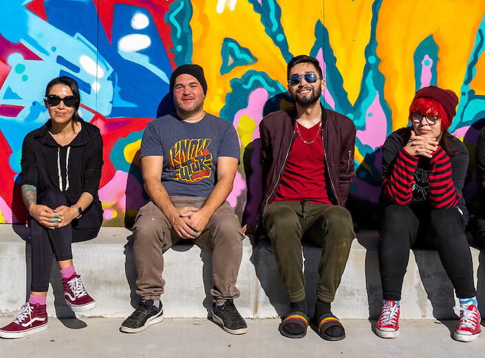 Mural Project. Mural project. Three students sit with instructor on curb in front of a bright mural. One female/slim/Latina in a black hoodie, one slim and white or Latino with green pants and red shirt, and the other female with bright red pink hair under a maroon beanie and striped red and black long-sleeved shirt. The instructor is in a t-shirt with a yellow/red logo (non discernible), a black beanie, and dark tan pants. He is Latino and in late twenties/thirties.