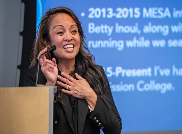 Speaker smiling and presenting at a podium with a projected slide showing her Mission College milestones.