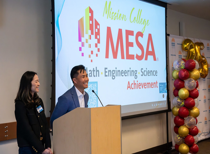 Two MESA program presenters speaking at a podium in front of a Mission College MESA banner and gold balloons.