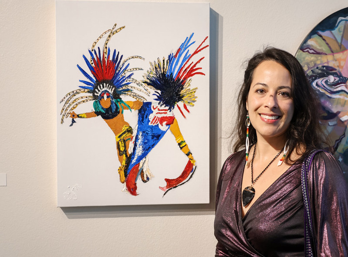 Artist standing beside a colorful painting of traditional dancers at the Mission College art exhibit.