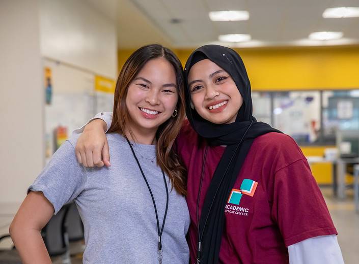 ASC Girls Two young women pose together in the Academic Success Center. One wears a hojab, the other is East Asian.