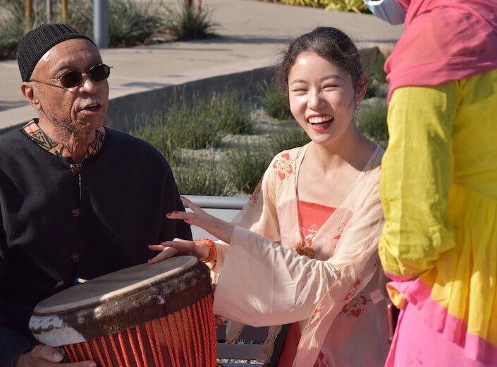 International Day International Day - a young woman of Asian descent in traditional Southeast Asian clothing drums on a Djembe belonging to Dr. Piper (an older man of African-American descene in traditional clothing.)