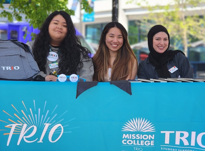 TRIO table. Three young women of color are sitting at TRIO booth on sunny day. One wears a jihab, the others have olive skin and long dark hair.